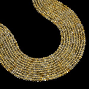 May include: A collection of strands of golden-yellow gemstone beads against a black background. The beads are small, rectangular, and translucent, with a slightly cloudy appearance. The beads are strung together, creating a flowing, curved shape.