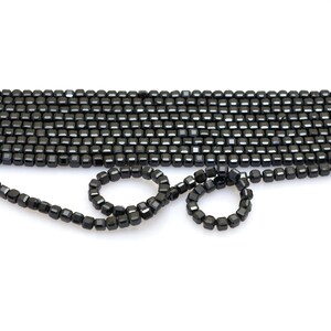 May include: Close-up of black, square-cut beads. The beads are highly reflective, with multiple facets catching the light. The beads are strung together, with some arranged in a curved pattern.