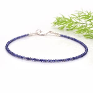 Può includere: Delicato braccialetto di perline blu con chiusura argentata. Il braccialetto è composto da piccole perline blu rotonde infilate insieme. La chiusura è argentata e assicura il braccialetto. Sfondo bianco con un tocco di verde.