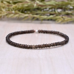 May include: A delicate bracelet featuring small, faceted, smoky quartz beads. The beads are a warm, earthy brown color and strung together with a silver clasp. The bracelet is displayed on a light-colored wooden surface, with a blurred green plant in the background.