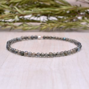 Puede incluir: Un brazalete delicado hecho con pequeñas cuentas redondas de labradorita iridiscente. El brazalete está enhebrado en una cadena de plata y tiene un diseño simple y minimalista.