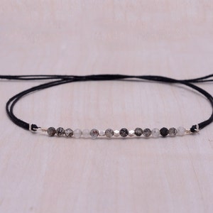 Op de afbeelding: Een armband van zwart koord met een zilveren sluiting en een rij grijze en zwarte edelsteen kralen.