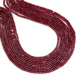 May include: Close-up of multiple strands of small, square-cut, deep red gemstones. The beads are strung together, creating a cascading effect. The gemstones have a slightly reflective surface, and the background is white.