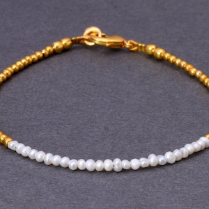 May include: Elegant gold bracelet with a strand of small, white pearls. The bracelet features a gold clasp and is accented with gold beads. The design is simple and elegant, suitable for everyday wear or special occasions.