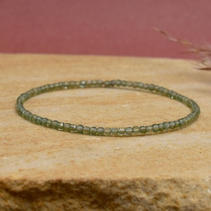 May include: A fine bracelet made of tiny, olive-green beads. The beads are threaded to create a flexible band. The bracelet is shown on a textured, light brown surface, with a soft, blurred background.