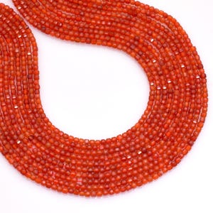 May include: Close-up of multiple strands of small, faceted, orange-red beads. The beads are strung together, creating a flowing, curved shape against a white background. The beads appear to be uniform in size and shape, suggesting a consistent design.