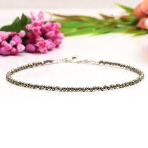 Könnte beinhalten: Ein filigranes Armband mit kleinen, runden, metallischen Perlen. Das Armband hat einen silberfarbenen Verschluss und wird auf einer weißen Oberfläche präsentiert. Die Perlen haben einen subtilen Schimmer, der auf einen Naturstein oder ein Mineral hindeutet.
