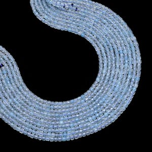 May include: Close-up of multiple strands of light blue square-cut beads. The beads are strung together, creating a flowing, curved shape against a black background. The beads have a subtle, translucent quality.