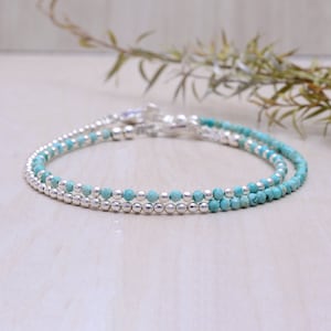 Peut inclure: Deux bracelets délicats avec des perles turquoise et de petites perles argentées. Les bracelets sont reliés par un fermoir argenté. Les bracelets sont présentés sur une surface de couleur claire.
