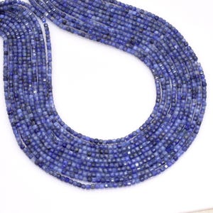 May include: A collection of strands of blue gemstone beads. The beads are small, square-cut, and have a faceted surface that reflects light. The color is a deep, rich blue with variations in tone. The beads are strung together, forming a curved shape.