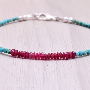 Könnte beinhalten: Ein zartes Armband mit kleinen, runden türkisfarbenen Perlen und einem zentralen Abschnitt aus leuchtend rot-rosa Edelsteinen. Das Armband wird mit einem silbernen Verschluss gesichert und ist ein schlichtes, aber elegantes Schmuckstück.