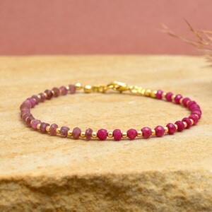 May include: A delicate bracelet featuring small, faceted, pink gemstones and gold-coloured beads. The bracelet is strung on a thin, gold-coloured chain and has a gold-coloured clasp. The jewellery is displayed on a light brown surface.