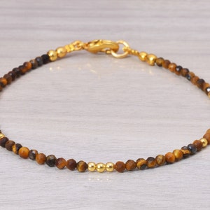May include: A gold and brown beaded bracelet with a clasp closure. The bracelet is made with tiger's eye and hematite beads.