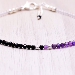 Puede incluir: Una delicada pulsera con un degradado de cuentas negras, moradas y blancas. La pulsera tiene un cierre plateado y pequeñas cuentas plateadas. Las cuentas son pequeñas y facetadas, creando un efecto brillante.