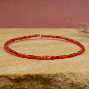 Puede incluir: Una delicada pulsera de cuentas de color rojo anaranjado. Las pequeñas cuentas cuadradas están ensartadas para formar una banda flexible. La pulsera se muestra sobre una superficie texturizada de color marrón claro, con un fondo suave y borroso.