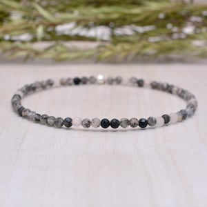 May include: A delicate bracelet made with small, round beads in shades of gray, black, and white. The bracelet is made with natural stones and has a simple, elegant design.