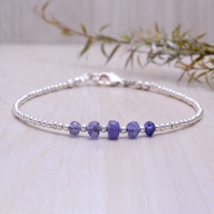 Puede incluir: Un delicado brazalete de plata con una fila de pequeñas piedras de amatista facetadas. El brazalete está hecho con pequeñas cuentas de plata y tiene un cierre simple.