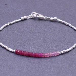 Può includere: Un braccialetto d'argento con una fila di perline di pietre preziose rosa e rosse. Il braccialetto è realizzato con piccole perline d'argento sfaccettate.