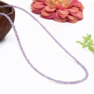 Könnte beinhalten: Eine zarte Halskette aus kleinen, hellvioletten Amethystperlen. Die Halskette wird auf einer weißen Oberfläche mit einer rosa Blume und grünem Laub im Hintergrund gezeigt.