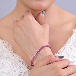 May include: A delicate bracelet of small, round, magenta beads. The bracelet is worn on a wrist, highlighting its vibrant colour and simple design. The beads appear to be strung together, creating a flexible and elegant accessory.