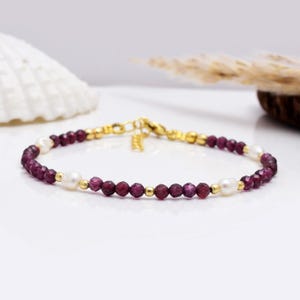 Könnte beinhalten: Ein zartes Armband mit facettierten, tiefroten Edelsteinen und glänzenden weißen Perlen. Das Armband ist mit goldfarbenen Perlen und einem goldfarbenen Verschluss versehen. Dieses elegante Schmuckstück eignet sich für den Alltag oder besondere Anlässe.