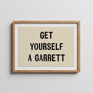 May include: A framed wall art print featuring a light beige background with the black text GET YOURSELF A GARRETT centred in a bold sans-serif font. The artwork is enclosed in a simple light brown wooden frame with a clean white mat border.