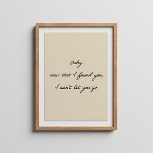 May include: A framed wall art print featuring a light beige background with black cursive text that reads: Baby, now that I found you, I can't let you go. The print is enclosed in a simple, light-coloured wooden frame with a clean, rectangular design.