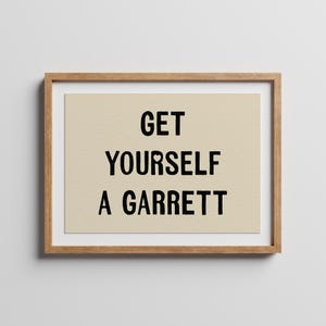 May include: A framed wall art print featuring a light beige background with the black text "GET YOURSELF A GARRETT" printed in a bold, sans-serif font. The artwork is enclosed in a simple, light-toned wooden frame with a clean, rectangular border.