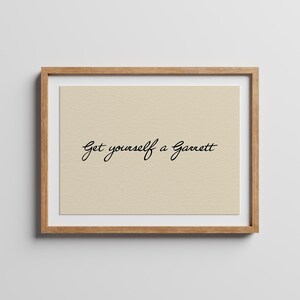 May include: A framed wall art piece featuring a light beige background with the black cursive text Get yourself a Garrett centred in the middle. The artwork is enclosed in a simple light brown wooden frame with a clean and neutral aesthetic.