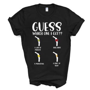 May include: Black t-shirt with white text that reads "GUESS WHICH ONE I GOT??".  The shirt features four illustrations of a leg with different items wrapped around it.  The items are a jar of pickles, BBQ ribs, a pickleball, and a pint of beer.