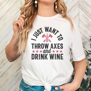 May include: White t-shirt with the text "I just want to throw axes and drink wine" in black with a pink wine bottle and crossed axes graphic.
