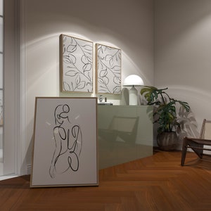 May include: A light green cabinet with two framed prints of black line drawings of leaves on a white background. A white lamp with a round shade sits on top of the cabinet. A brown wooden chair with a woven seat sits in the corner of the room. A large framed print of a black line drawing of a person sitting with their back to the viewer is on the floor in front of the cabinet.
