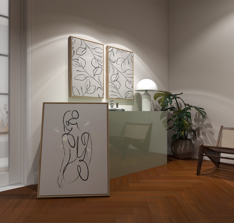May include: A light green cabinet with two framed prints of black line drawings of leaves on a white background. A white lamp with a round shade sits on top of the cabinet. A brown wooden chair with a woven seat sits in the corner of the room. A large framed print of a black line drawing of a person sitting with their back to the viewer is on the floor in front of the cabinet.