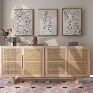 May include: A light wood cabinet with four doors and a woven rattan front. The cabinet is sitting on a pink and black tiled floor. Three framed black and white line drawings are hanging on the wall above the cabinet.