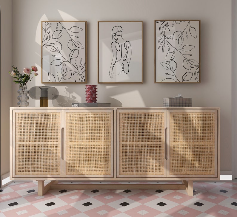 May include: A light wood cabinet with four doors and a woven rattan front. The cabinet is sitting on a pink and black tiled floor. Three framed black and white line drawings are hanging on the wall above the cabinet.