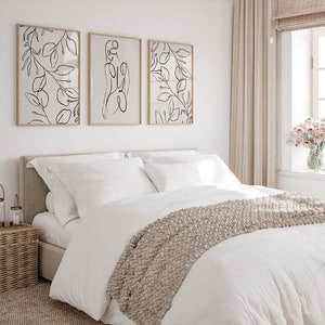 May include: A white bed with a beige knitted throw blanket and white pillows. Three framed black and white line drawings of leaves and a woman's figure hang on the wall above the bed.