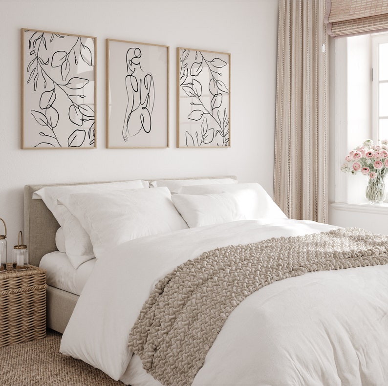 May include: A white bed with a beige knitted throw blanket and white pillows. Three framed black and white line drawings of leaves and a woman's figure hang on the wall above the bed.
