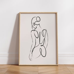 May include: A black and white line drawing of a person's back, in a simple, minimalist style. The drawing is framed in a light wood frame and is on a light wood floor.