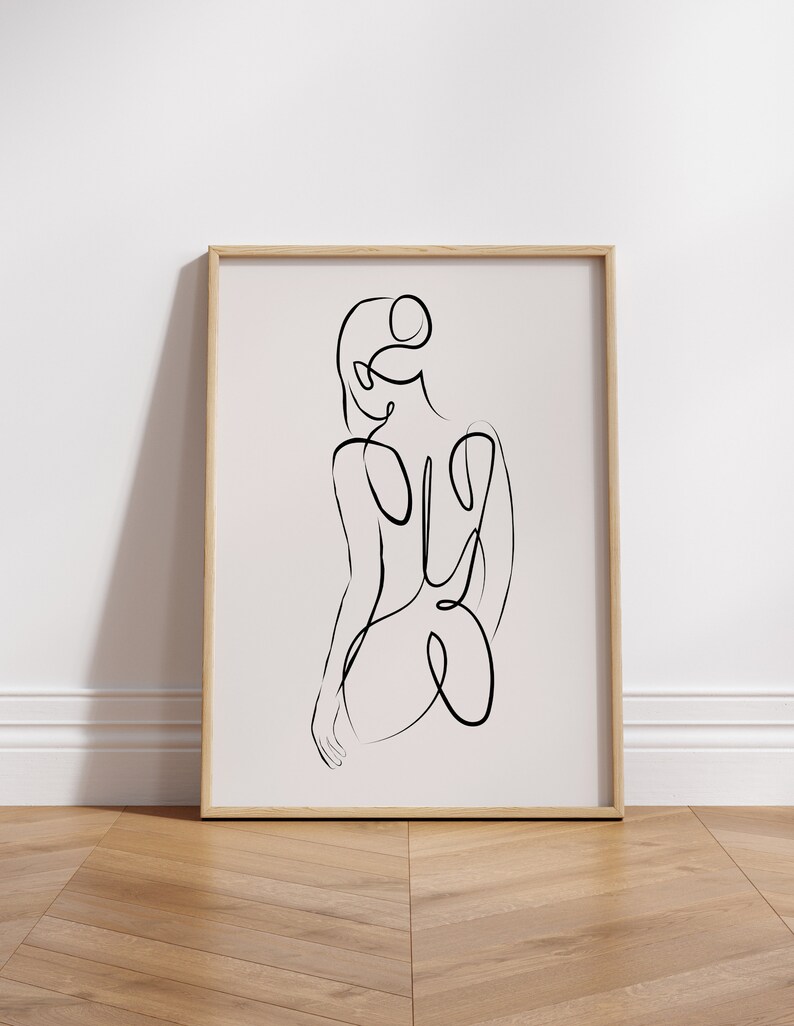 May include: A black and white line drawing of a person's back, in a simple, minimalist style. The drawing is framed in a light wood frame and is on a light wood floor.