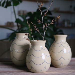 Könnte beinhalten: Drei Keramikvasen mit cremefarbener Glasur und blauen Wellenlinien. Die Vasen sind in einer Reihe auf einer Holzoberfläche angeordnet. Ein Zweig mit braunen Knospen befindet sich in der mittleren Vase.