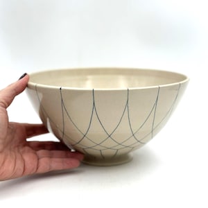 May include: Cream-colored ceramic bowl with a pedestal base. The bowl features a delicate pattern of intersecting navy blue lines. The bowl's design is simple and elegant, suitable for serving or display.