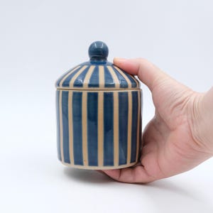 Puede incluir: Un tarro cilíndrico de cerámica con tapa abovedada a juego, que presenta un patrón de rayas verticales repetidas en azul oscuro y beige claro. El recipiente tiene un acabado liso y brillante con un pequeño pomo redondo en la tapa.