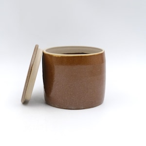 May include: A brown ceramic jar with a lid resting beside it. The jar has a cylindrical shape with a slightly textured surface and a lighter brown rim. The lid is a flat disc with a similar color scheme.
