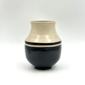 Puede incluir: Un jarrón de cerámica con la parte superior color crema y la base negra. Una franja negra rodea el jarrón donde se encuentran los colores. El jarrón tiene forma redondeada y una abertura ancha, adecuado para exhibir flores o como pieza decorativa.