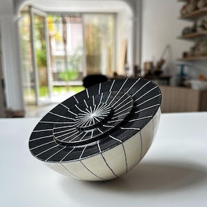 Op de afbeelding: Een gehalveerde keramische kom met een uniek ontwerp. De kom onthult drie geneste, cirkelvormige platforms. De platforms en het buitenoppervlak zijn zwart met witte lineaire patronen. De basis is gebroken wit.
