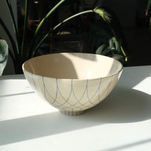 Könnte beinhalten: Eine cremefarbene Keramikschale mit blauem geometrischen Muster. Die Schale hat eine breite, abgerundete Form und einen Fuß. Das blaue Muster besteht aus sich kreuzenden, gebogenen Linien. Die Schale steht auf einer weißen Oberfläche.