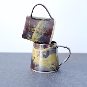 Può includere: Due tazze in ceramica impilate verticalmente, caratterizzate da una smaltatura di base marrone scuro lucido con accenti testurizzati gialli e dorati. Ogni tazza presenta un manico curvo liscio e un corpo cilindrico affusolato con bordo chiaro.