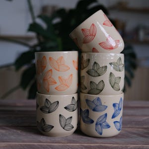 Può includere: Quattro tazze in ceramica impilate l'una sull'altra. Le tazze sono bianche con un motivo di foglie stilizzate in arancione, verde, blu e nero.