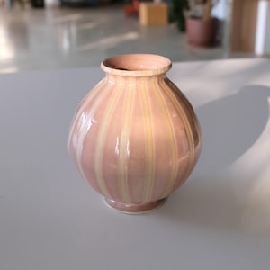 May include: A small, round, pink ceramic vase with vertical cream-colored stripes. The vase has a narrow neck and a slightly flared rim. The base is a small, circular foot. The vase is sitting on a white surface.