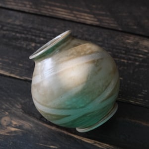 May include: A handmade ceramic vase with a rounded body and a slightly flared rim. The vase features a swirl pattern of tan and cream colors, with a touch of green at the base. The vase is approximately 5 inches tall.